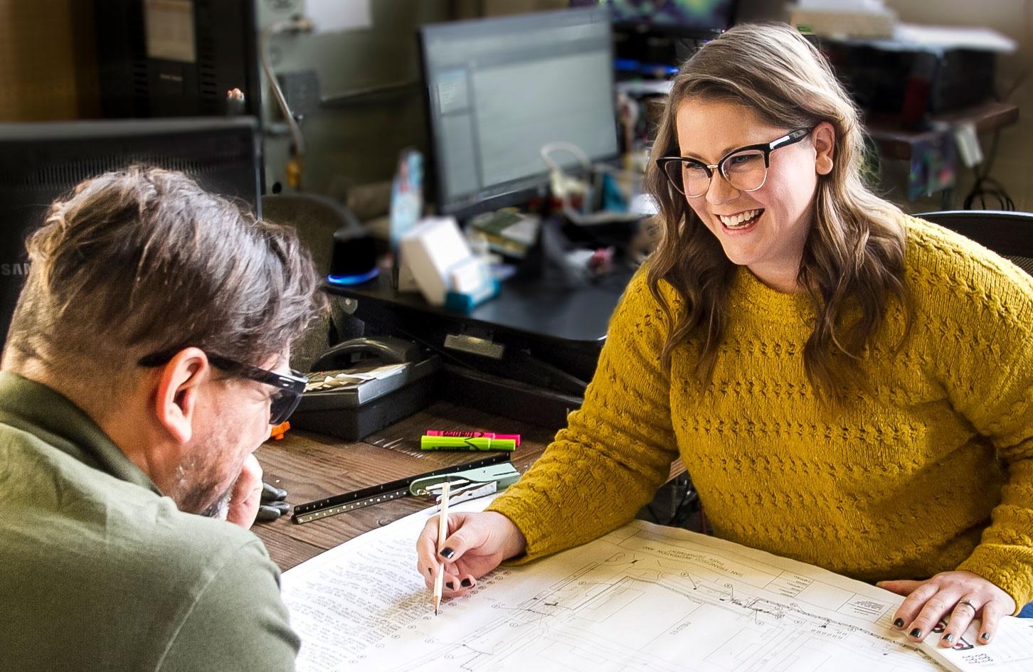 Person at a desk covered in plans and papers helping another person
